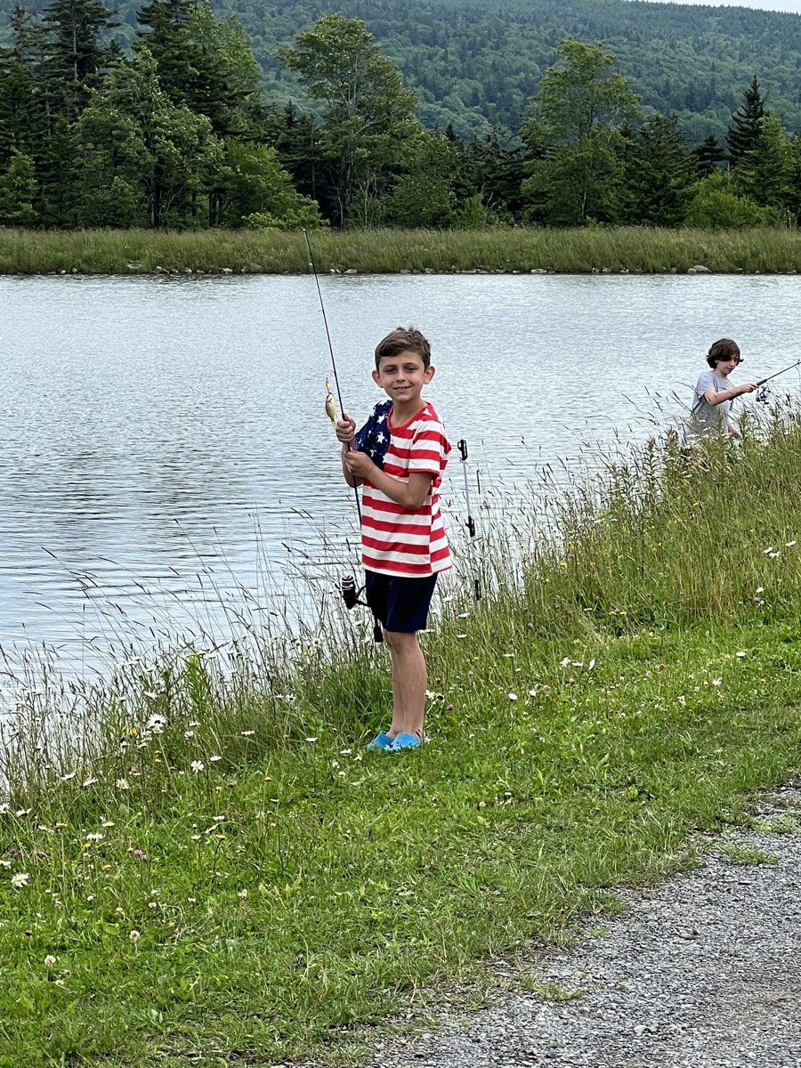 The boy in the flag t-shirt standing on the grassy bank with his rod bent and a small fish on the line, another child fishing further along the shore.