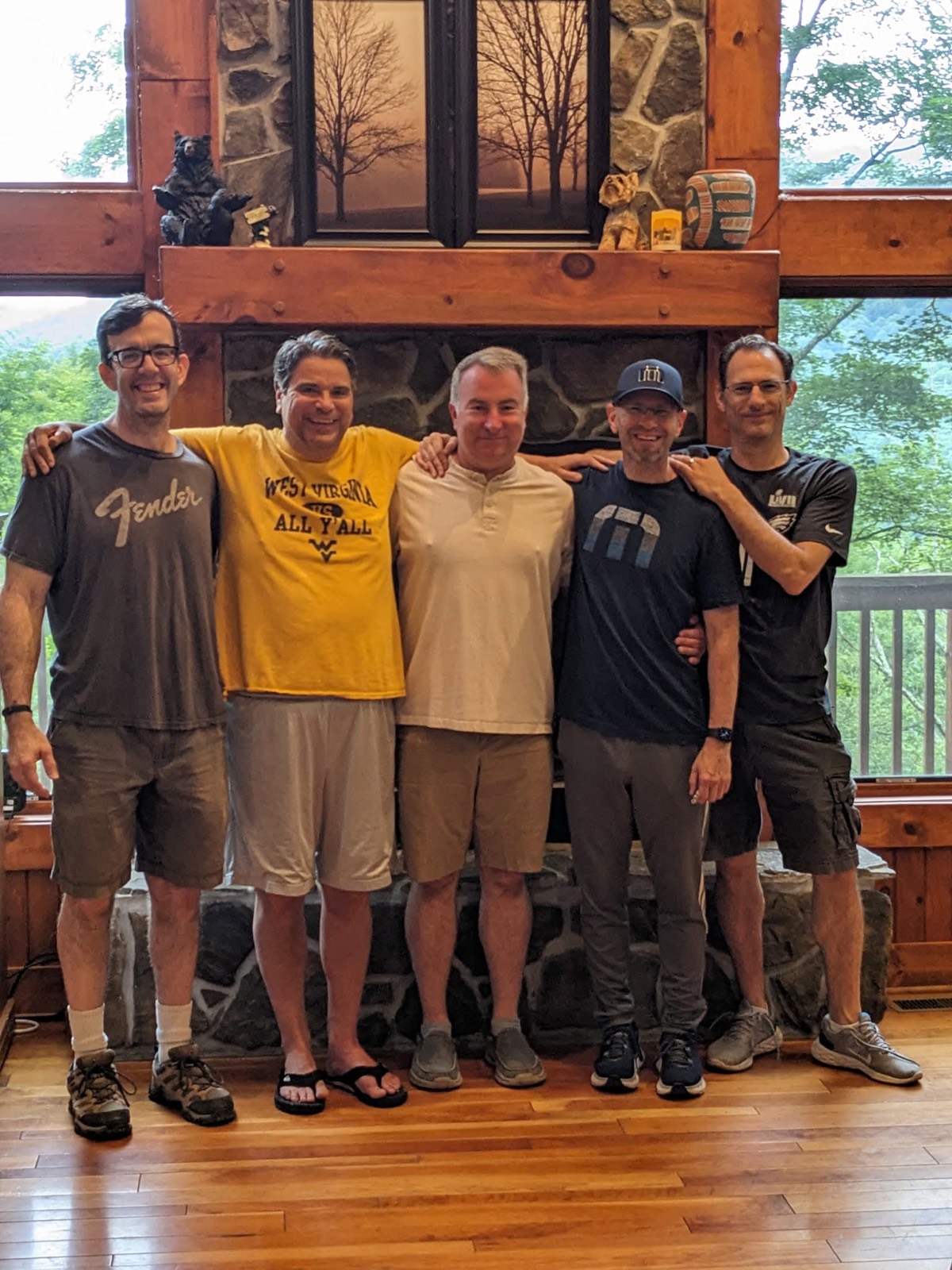 Five men standing shoulder to shoulder in front of a tall stone fireplace in the cabin's great room, arms around each other, big windows and a wooded deck behind them.