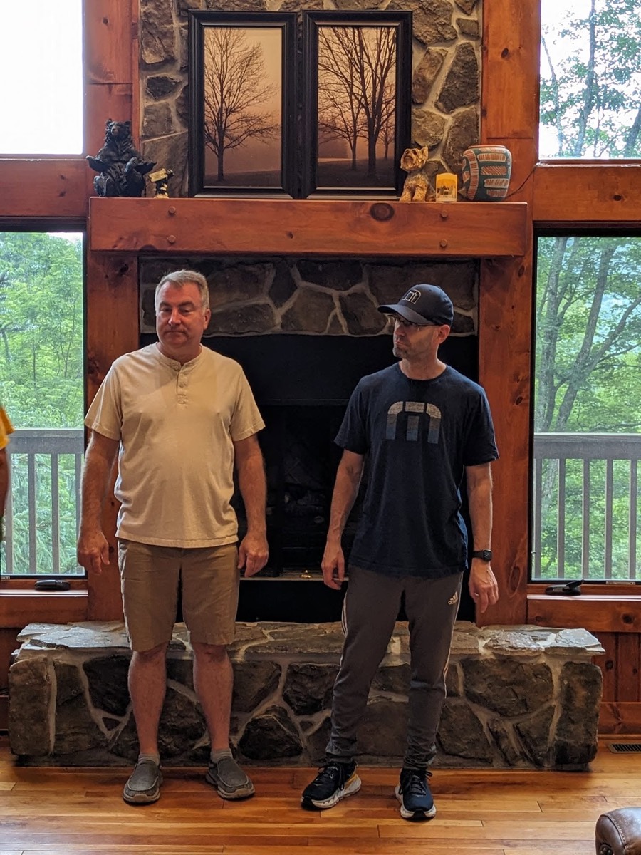 Two men standing on the raised stone hearth in front of the same fireplace, one in a tan shirt and one in a black t-shirt and cap.