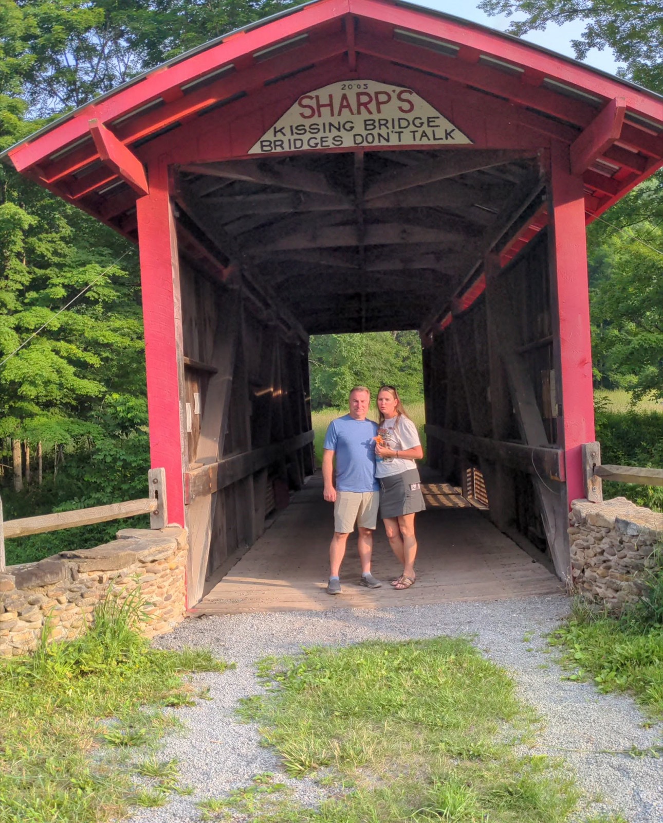 A red covered bridge photographed head-on from the road, its gable sign reading 2005, SHARP'S KISSING BRIDGE, BRIDGES DON'T TALK, with two people standing small in the dark opening of the span.