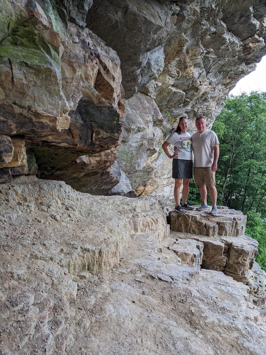 A huge tilted rock overhang of layered gray stone with two adults standing together on a ledge beneath it, green trees beyond.