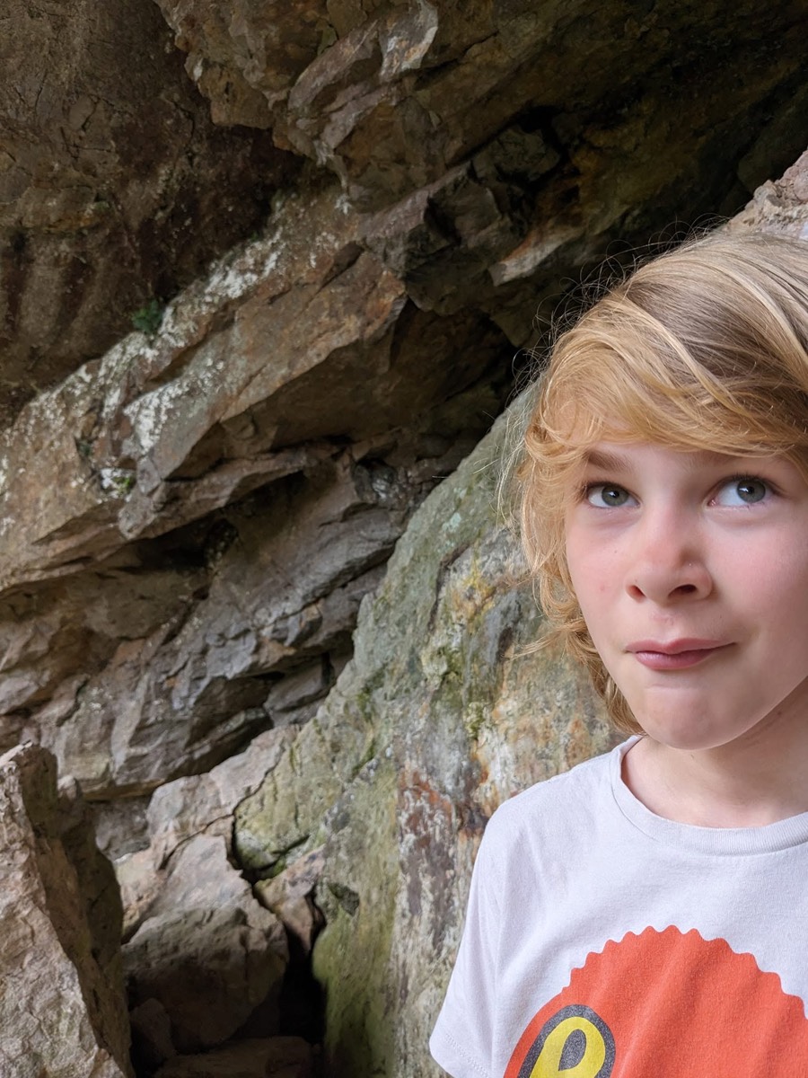 A blond child in a white t-shirt standing close against a mossy rock wall, looking off to the side, weathered stone filling the rest of the frame.