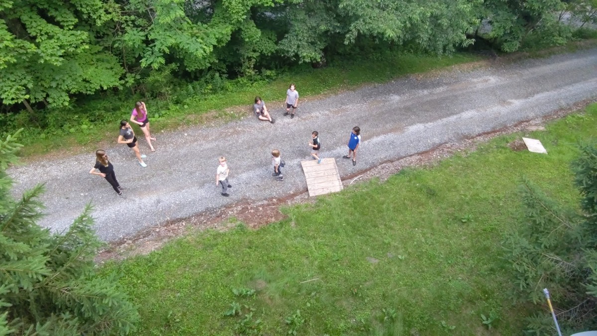 An aerial view looking down on a gravel lane running beside a mown green lawn and dense woods, with several small figures walking and sitting along it.