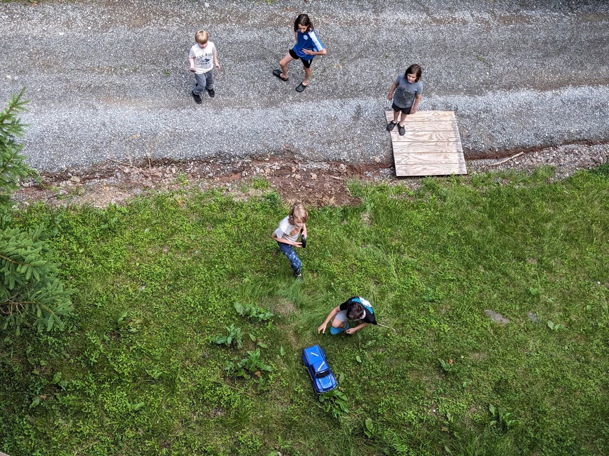 A near-vertical aerial view of a gravel lane and grass verge with five people walking across it, long shadows and a bright green lawn.