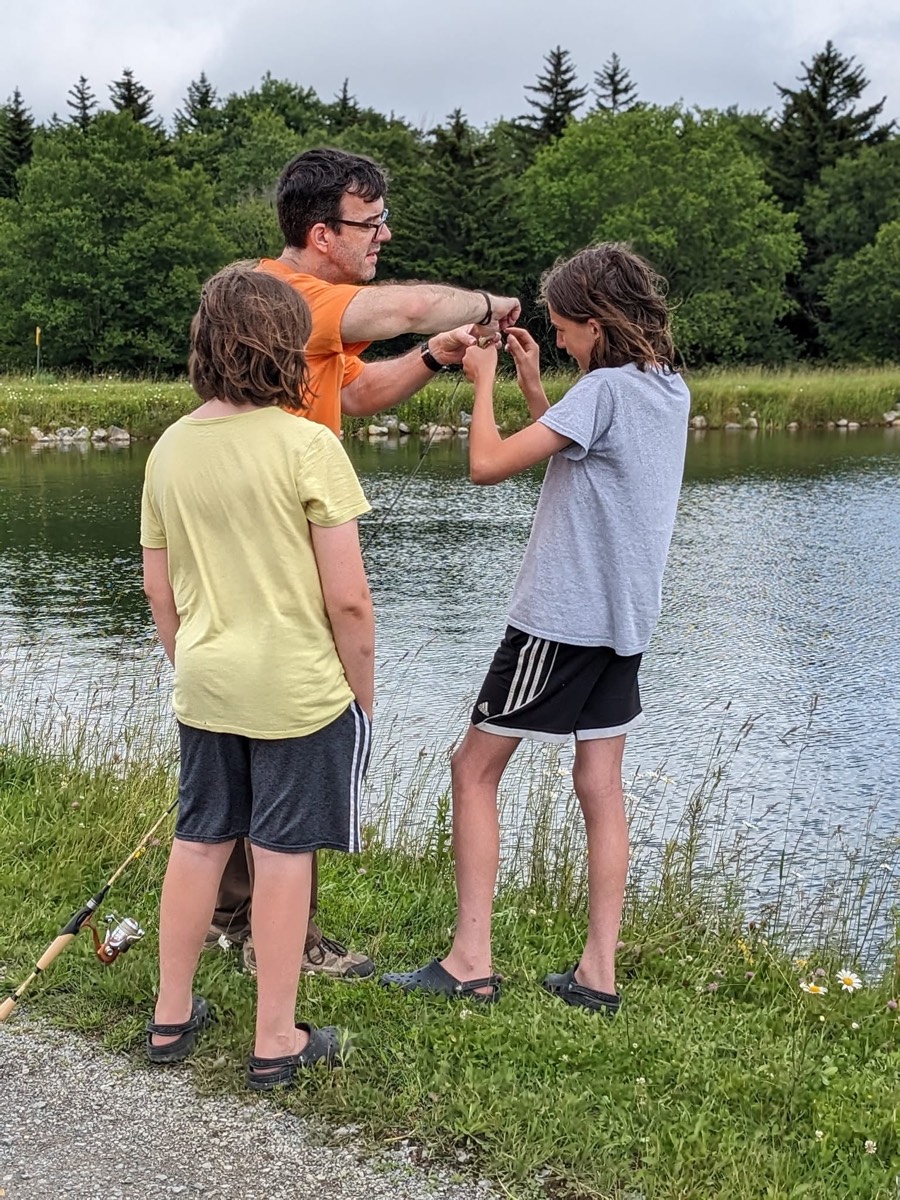 A man helping two children rig a fishing rod on the grass bank of the pond, dark spruce woods across the water behind them.