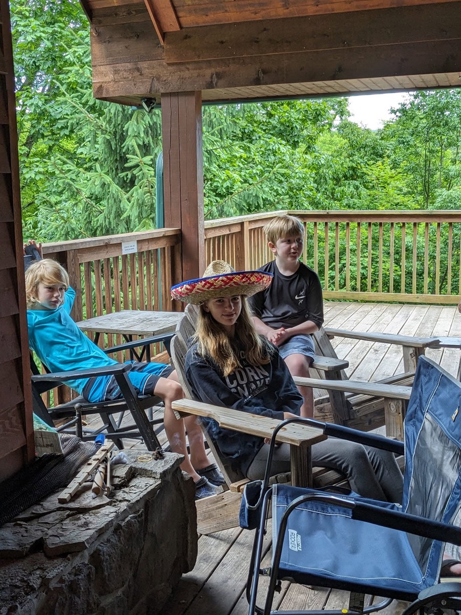 Several children and adults sitting in folding camp chairs on the cabin's covered porch, one wearing a straw sombrero, green woods beyond the railing.