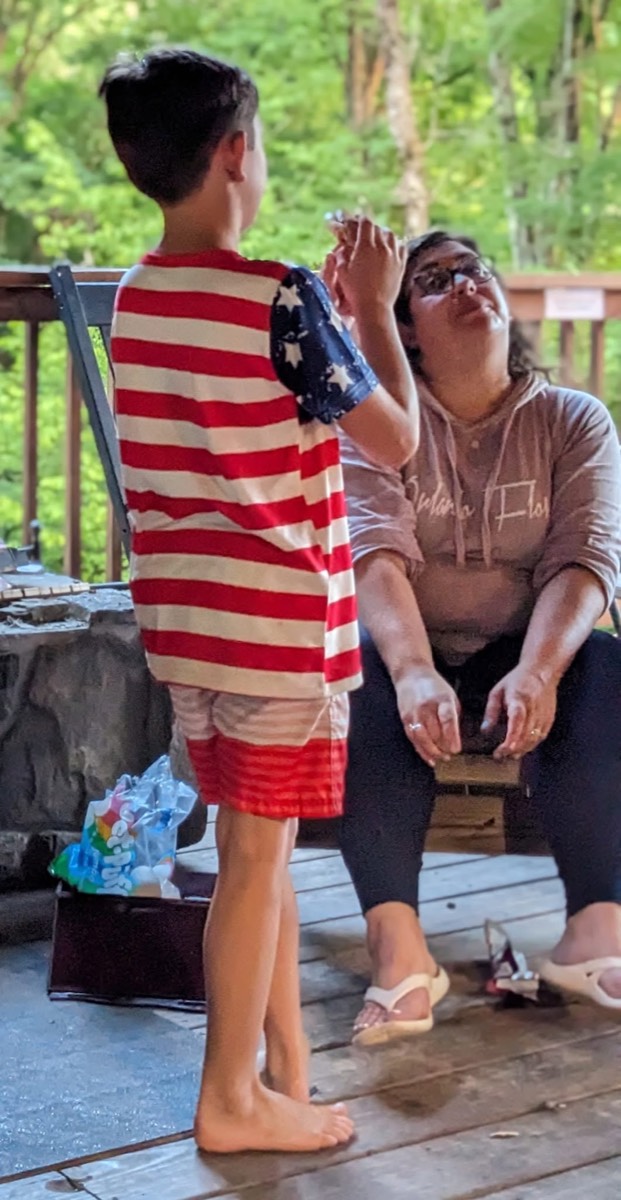 A boy in a stars-and-stripes shirt standing on the wet deck in front of a woman sitting on the stone step, both laughing.