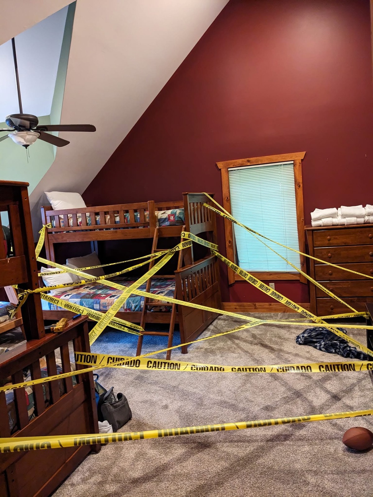 A cabin bunk room with deep red walls, its wooden bunk beds crisscrossed floor to ceiling with yellow CAUTION tape, a football on the carpet.