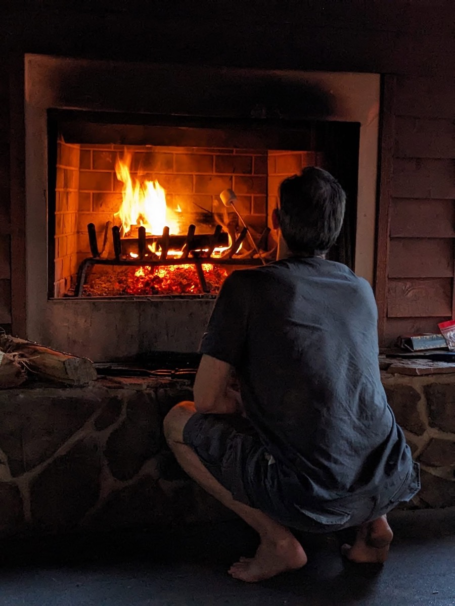 A person crouched in front of a large open fireplace with a substantial fire burning, seen from behind in silhouette against the flames.
