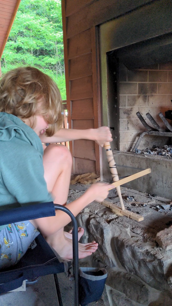 A child sitting in a camp chair holding a long wooden stick into the mouth of an outdoor stone fireplace on the porch.