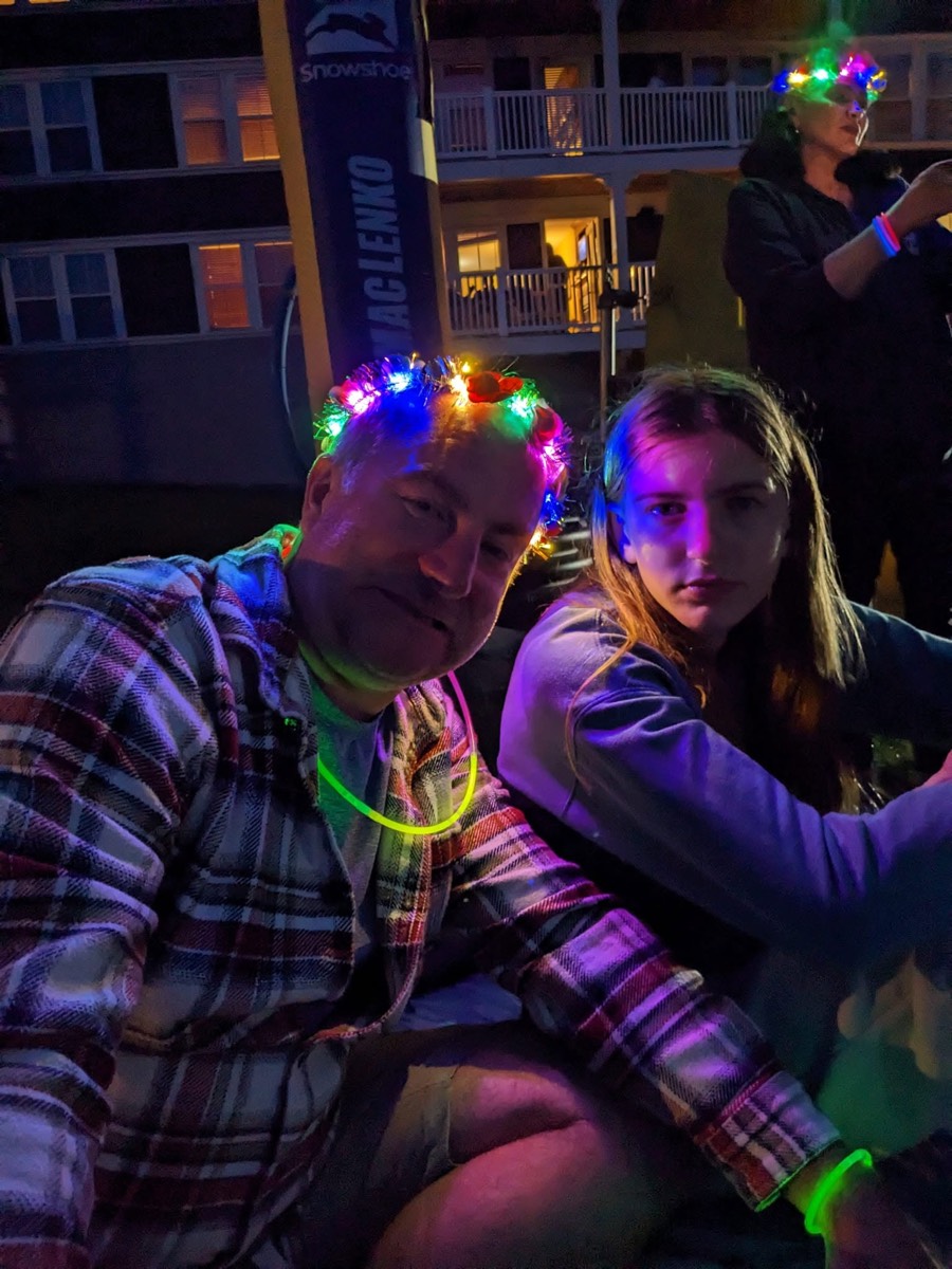 Two people sitting close together at night lit in purple and green by glow sticks and light-up crowns, a lit resort building and a Demaclenko banner behind them.
