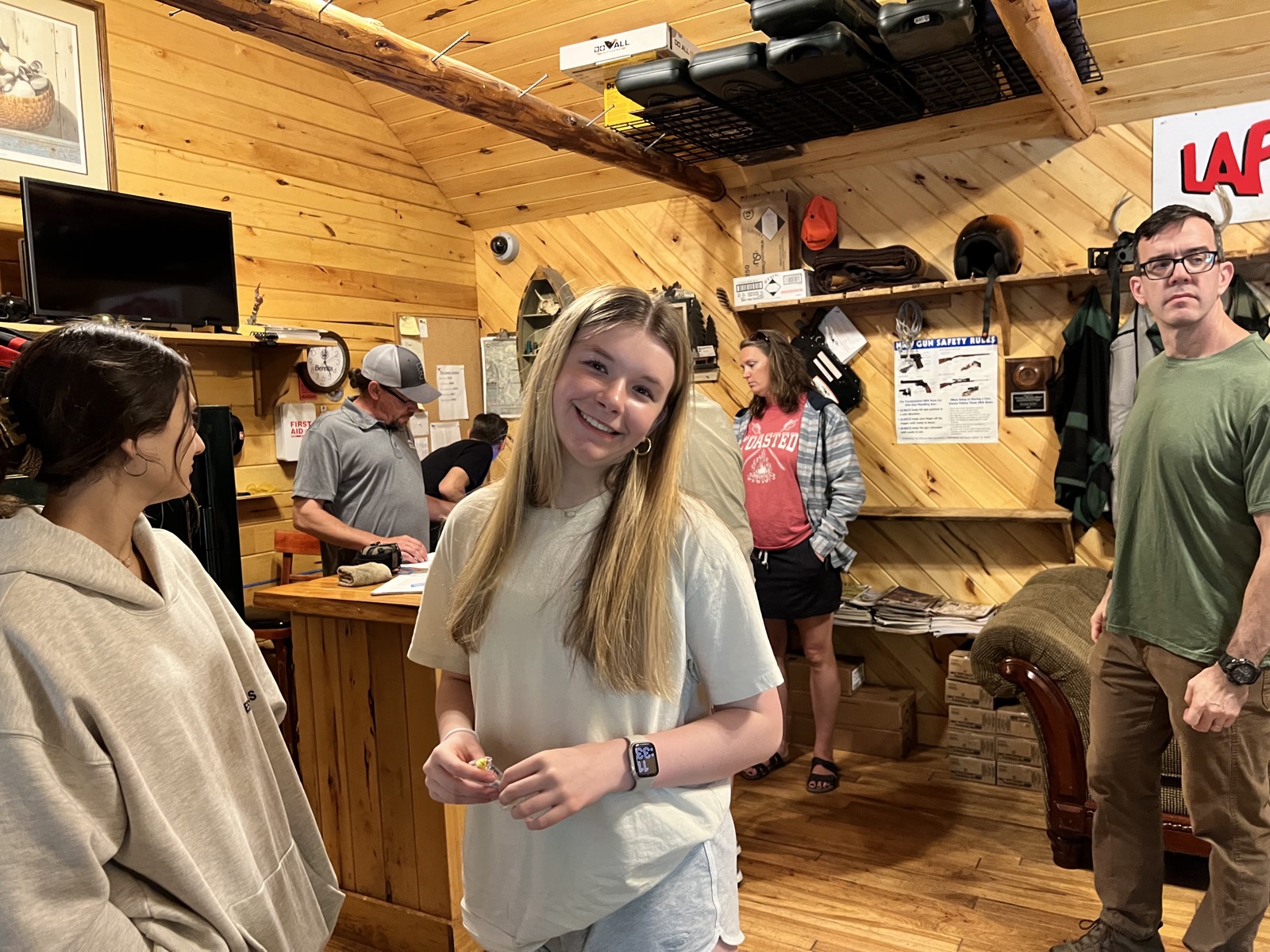 Inside a wood-panelled range clubhouse, a smiling girl in a pale t-shirt stands holding something small while others check in at a counter, a gun safety rules poster and boxes of ammunition on the shelves behind.