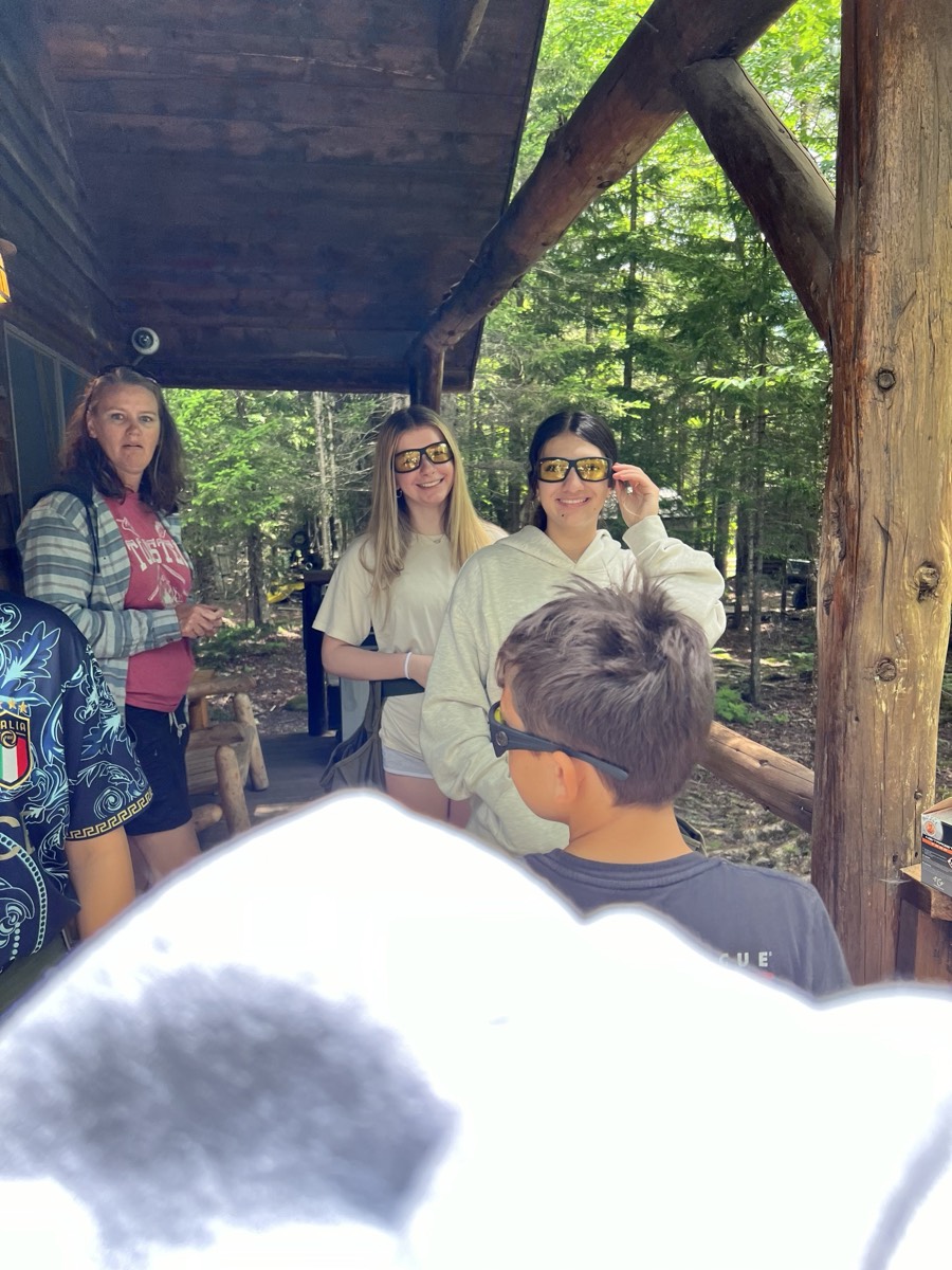 A group standing under a timber shooting stand wearing yellow-tinted safety glasses, green woods behind them.