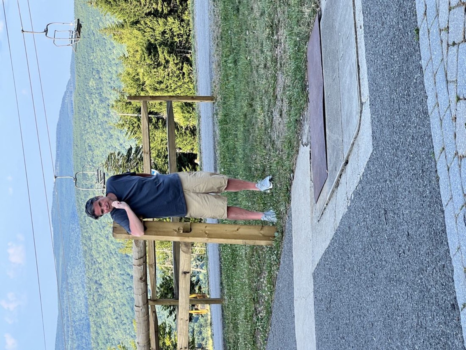 George leaning back against a wooden fence post with his fist under his chin in a thinker pose, empty ski lift chairs strung across the green mountain behind him at Snowshoe.