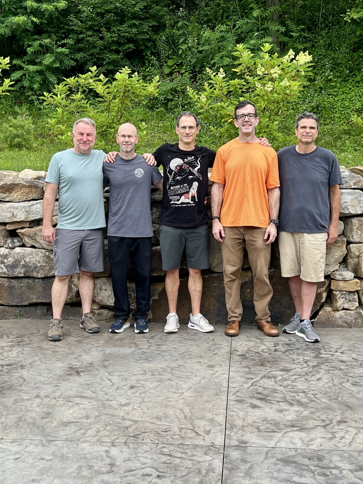 Five men standing shoulder to shoulder in front of a dry stone retaining wall with green woods and flowering shrubs behind them, arms around each other's shoulders. Left to right: Heath in a pale teal shirt, Shawn in a gray tee, Brian in a black Return of the Mothman t-shirt, Jim in an orange shirt, and George in a dark gray shirt.