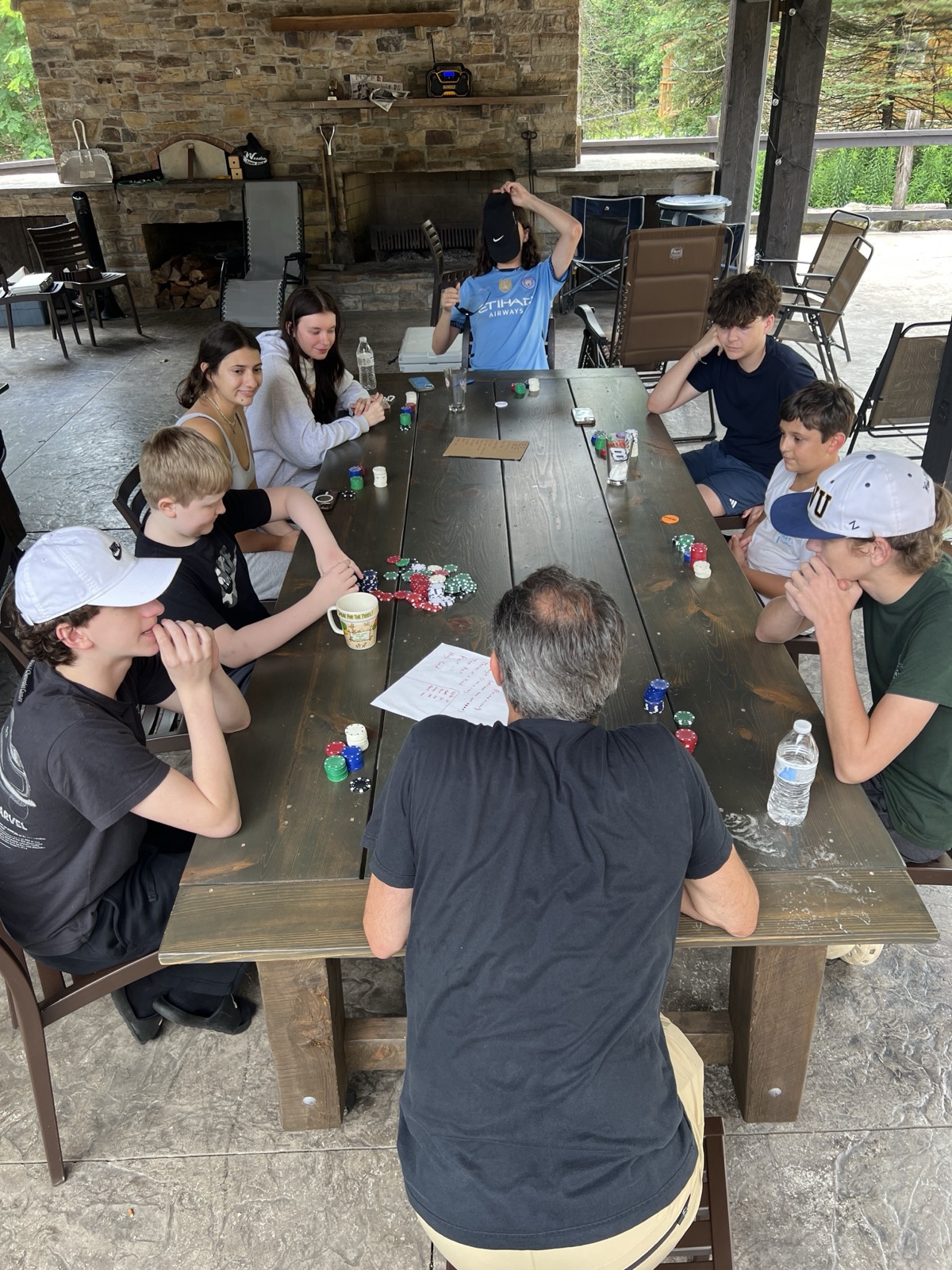 An overhead view of ten people, mostly teenagers, seated around a long dark wooden table on a covered stone patio. Poker chips are piled in the middle and a handwritten hand-rankings sheet lies on the table. A stone fireplace and stacked firewood behind them.