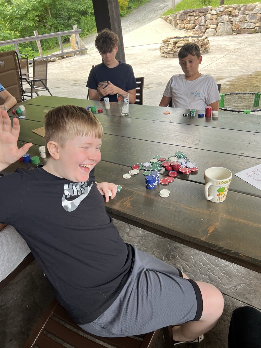 Owen Nardella, a grinning blond boy in a black Nike t-shirt, sitting at the poker table with one hand raised, reaching toward a large pile of chips in front of him while two other boys watch from across the table.