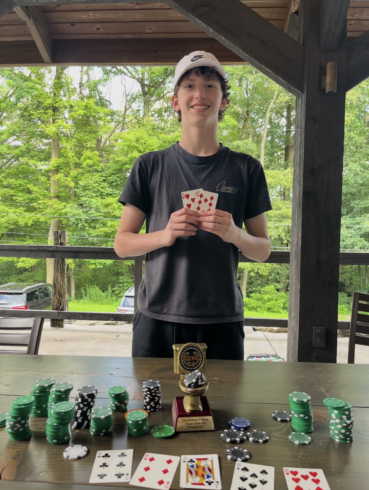 A teenage boy in a white Nike cap and dark t-shirt smiling behind a gold poker trophy engraved Chuckwagon Gang Texas Hold em Champion, holding up two playing cards, with tall stacks of green and black chips and five community cards spread across the table in front of him.