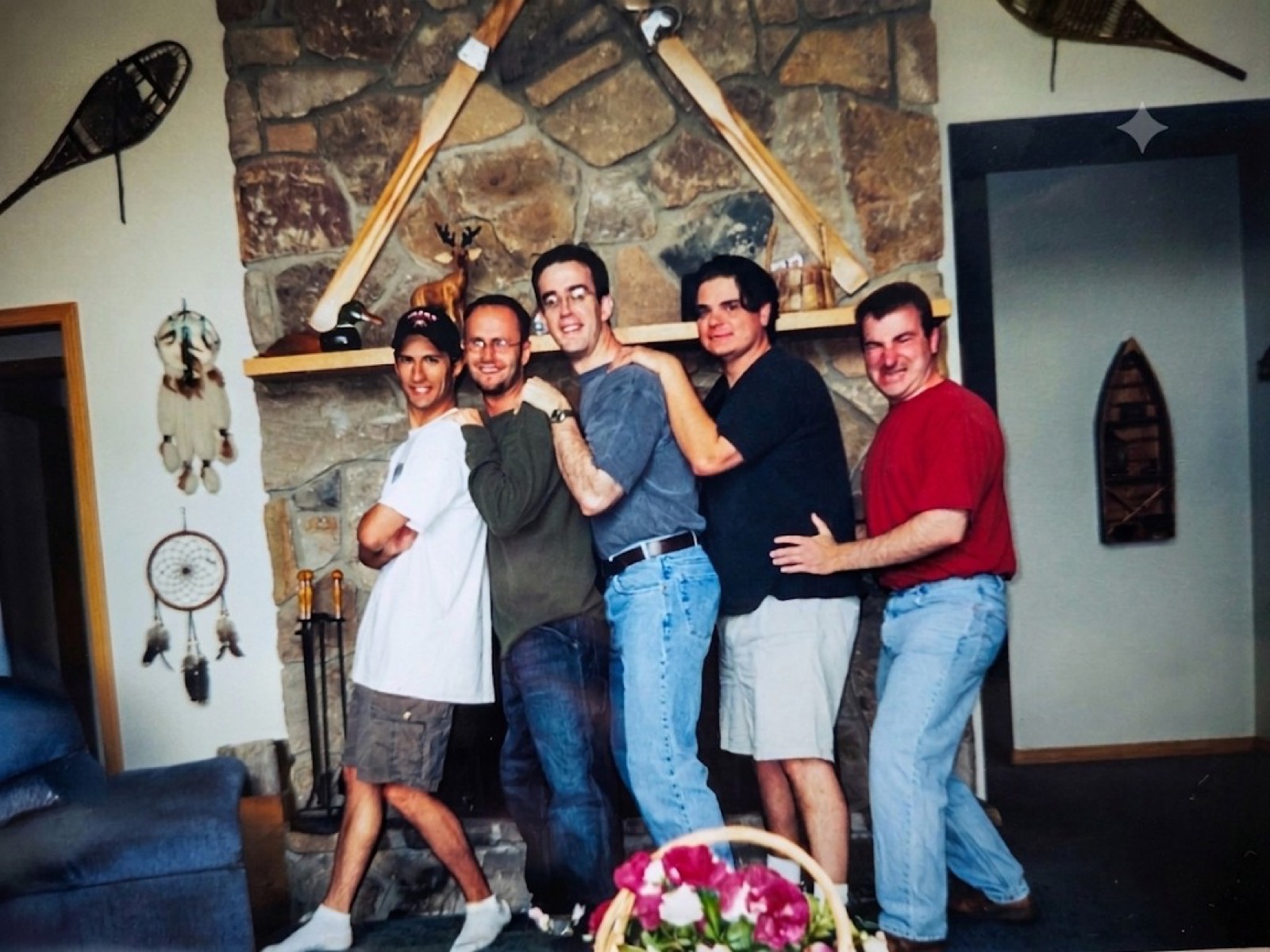 Five men standing in a staggered line in front of a tall stone fireplace, each resting a hand on the shoulder of the man ahead. Crossed wooden canoe paddles hang over the mantel, snowshoes and a dreamcatcher hang on the walls, and a basket of pink flowers sits in the foreground.