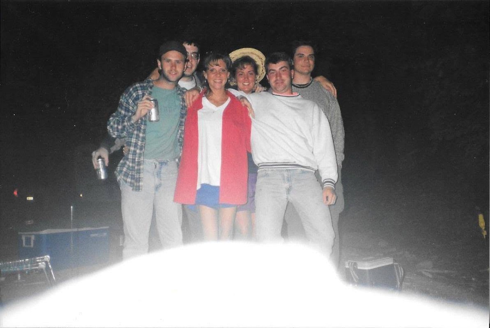 A night flash photograph of six people at a campsite. Left to right: a man in a plaid shirt and cap holding a can, a man in glasses standing behind him, a woman in a red jacket over a white top at the center front, a woman in a straw hat behind her, a man in a white sweatshirt, and a man in a gray sweater at the right edge.