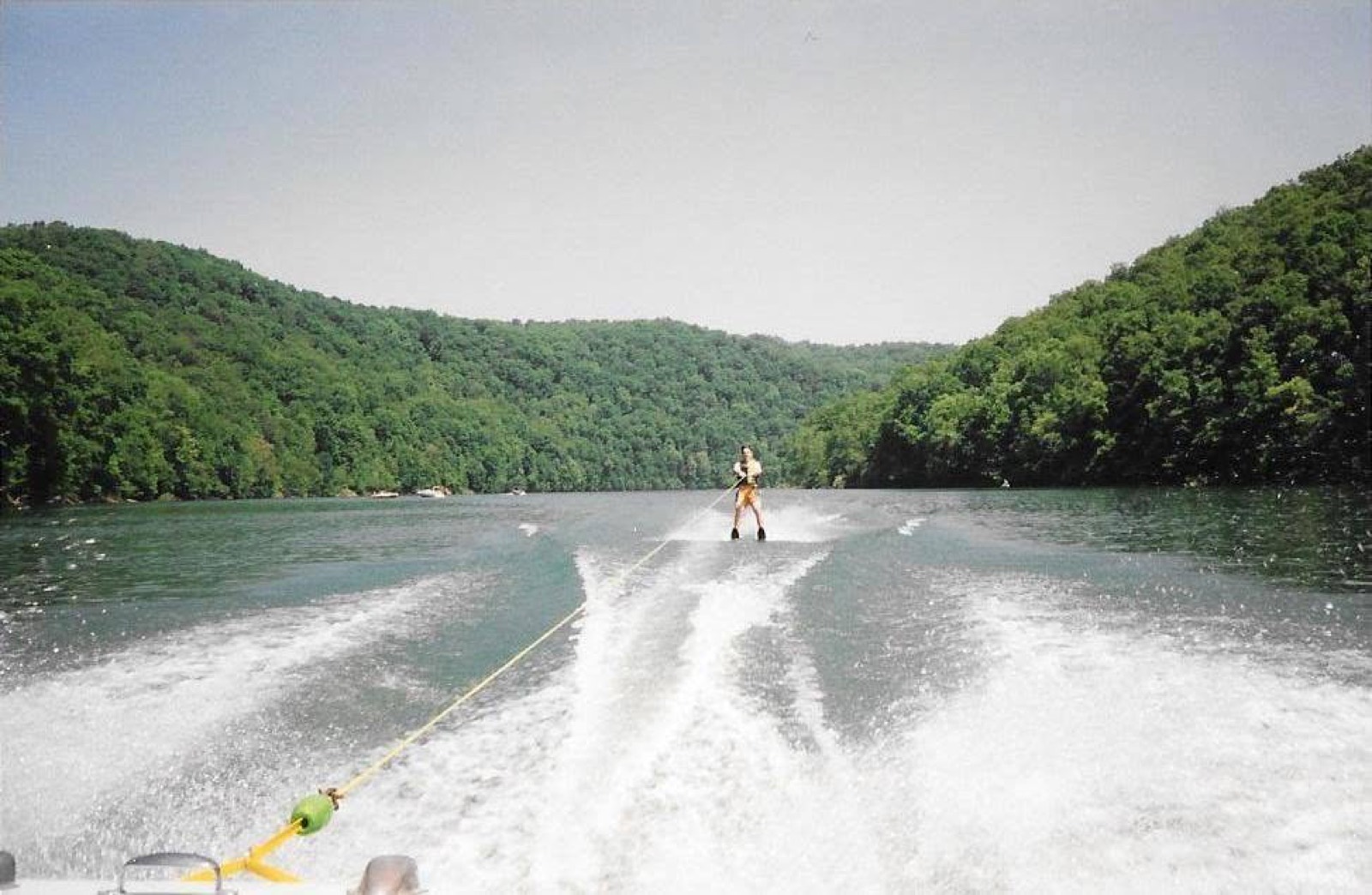 A water skier photographed from the back of a moving boat, riding the wake down a narrow stretch of green water between steep forested hills.