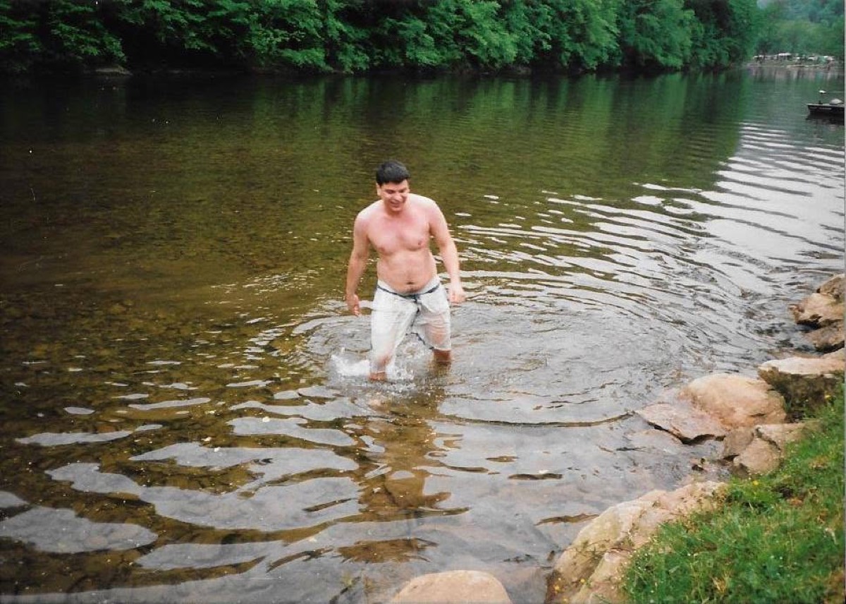 A man in white shorts wading out of a shallow green river toward a rocky bank, dense summer woods along the far shore.