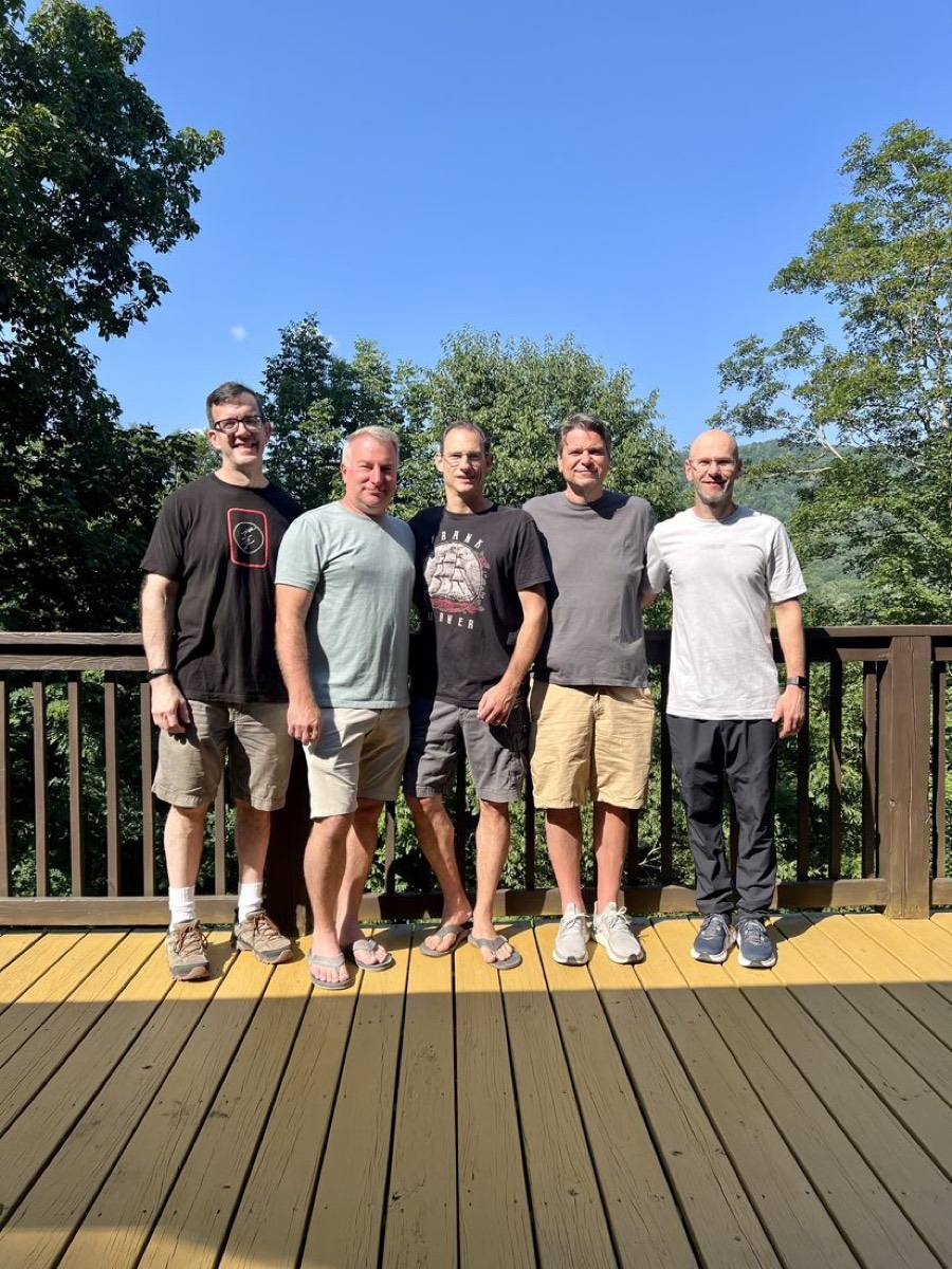 The five founders standing together on the cabin deck, blue sky and mountains behind them